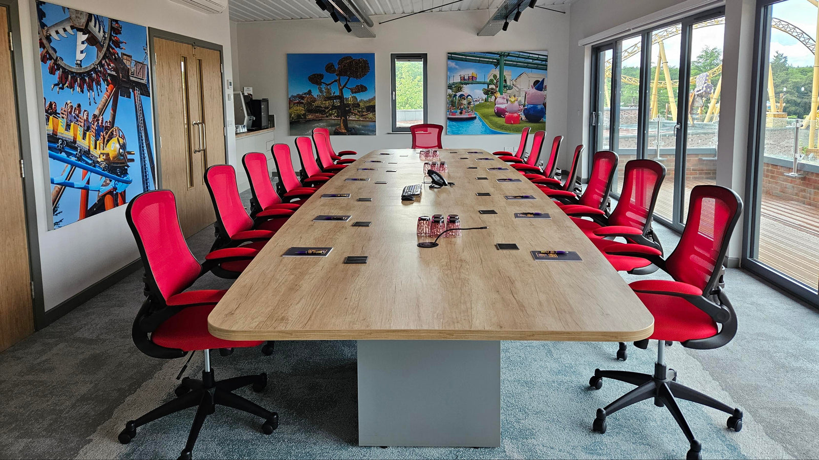 A modern conference room with a long light wood meeting table, red office chairs, large windows, and colorful wall murals depicting roller coasters and landscapes.
